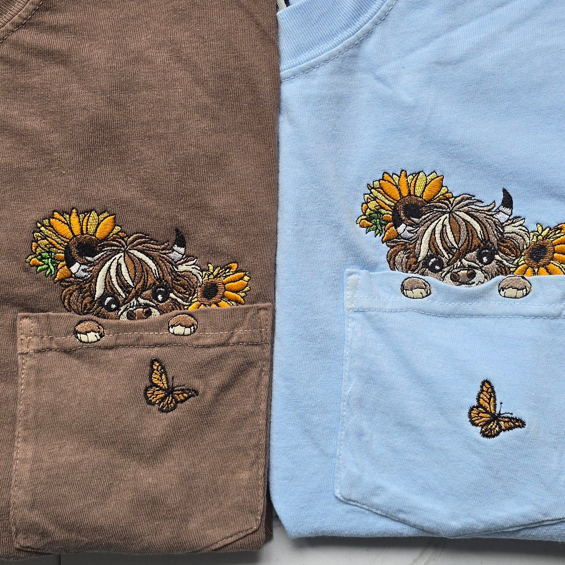 Two folded shirts, one brown and one blue, with embroidered designs of a highland cow and sunflowers.