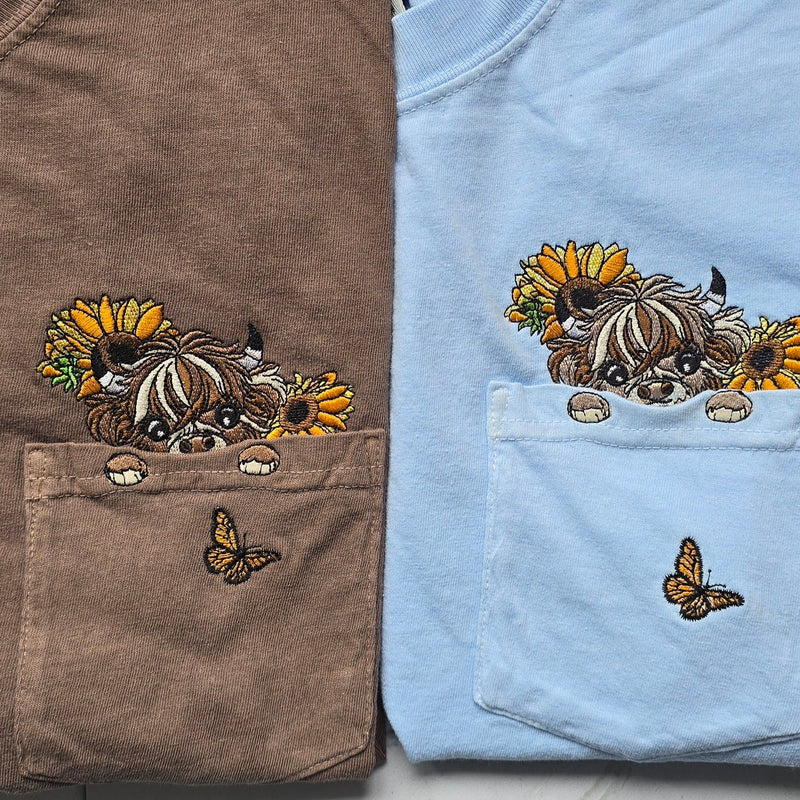 Two folded shirts, one brown and one blue, with embroidered designs of a highland cow and sunflowers.